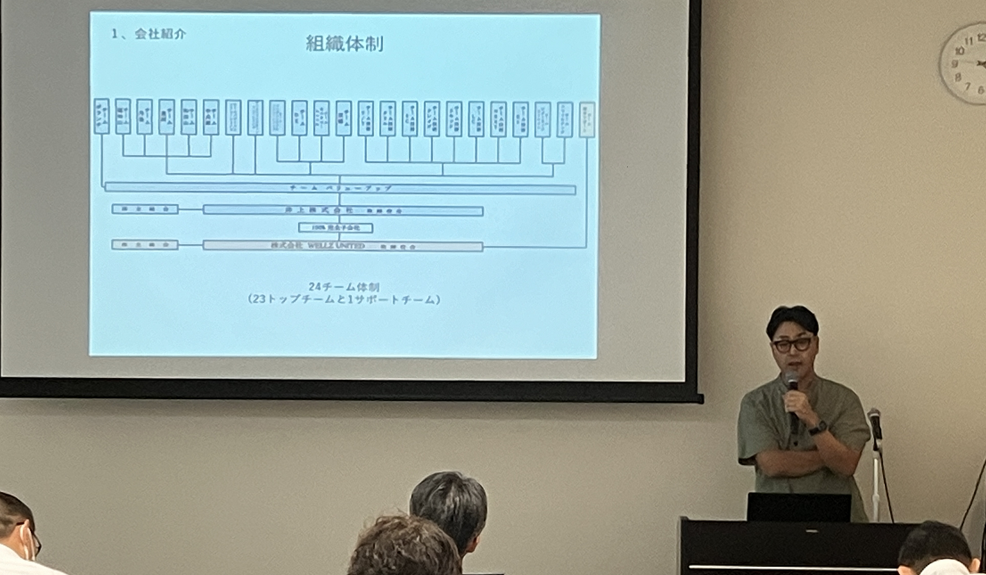  株式会社WELLZ UNITED代表取締役 井上氏による講演の様子。 同社の組織図は、経営側が「経営をサポートする」ことを表現するため、逆ピラミッド型となっている。